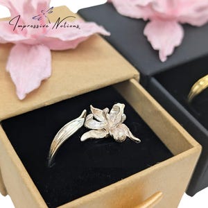 Puede incluir: Un anillo de plata con un delicado diseño floral. El anillo se muestra en una pequeña caja marrón con un forro de terciopelo negro. La caja está abierta y el anillo es visible en el centro de la caja.