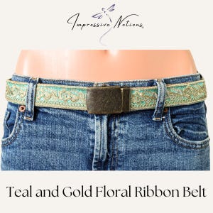 Puede incluir: Cinturón de cinta floral azul verdoso y dorado con una hebilla de metal marrón. El cinturón se muestra en un par de pantalones vaqueros azules.