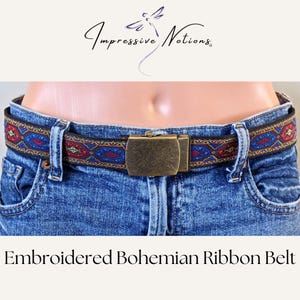 May include: A colorful embroidered ribbon belt with a geometric pattern in shades of red, blue, and gold. The belt has a brass buckle and is shown worn on a pair of blue jeans.