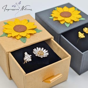 Puede incluir: Un par de pendientes de plata en una pequeña caja de cajones. Los pendientes son una margarita y una abeja. La caja está decorada con un girasol de papel y las palabras "Impressive Notions".