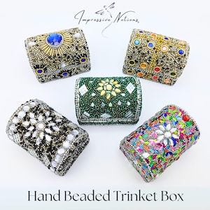 May include: Five small, hand-beaded trinket boxes in various colors and patterns. The boxes are made of metal and have a hinged lid. The boxes are decorated with beads, sequins, and rhinestones. The boxes are arranged in a circle on a white background. The text "Impressive Notions" is printed above the boxes. The text "Hand Beaded Trinket Box" is printed below the boxes.