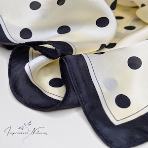 Puede incluir: Pañuelo de seda color crema con lunares negros grandes y borde negro. El pañuelo está doblado, mostrando su textura suave y diseño. Un accesorio de moda clásico.