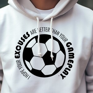 Puede incluir: Sudadera con capucha blanca con cordón. La parte delantera presenta un gráfico de balón de fútbol en blanco y negro con el texto: "I HOPE YOUR EXCUSES ARE BETTER THAN YOUR GAMEPLAY."