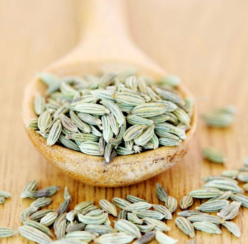 May include: A wooden spoon filled with green fennel seeds. The seeds are scattered on a wooden surface.