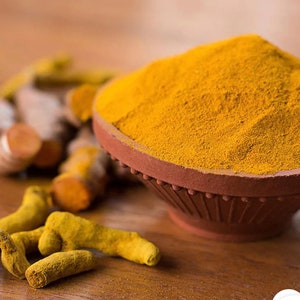 May include: A close-up of a bowl of bright yellow turmeric powder, with fresh turmeric root pieces in the foreground. The bowl is made of terracotta and has a decorative rim.