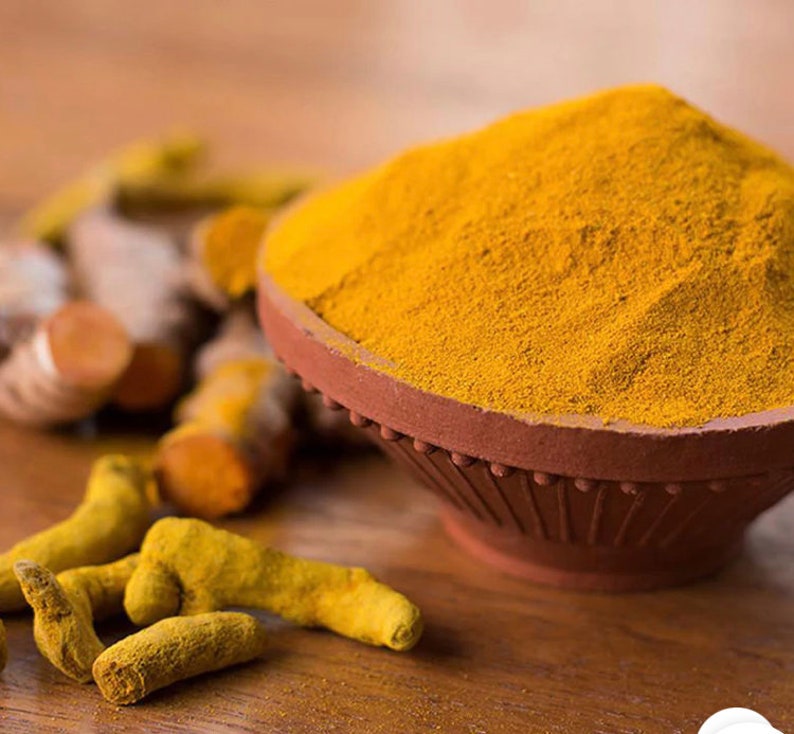 May include: A close-up of a bowl of bright yellow turmeric powder, with fresh turmeric root pieces in the foreground. The bowl is made of terracotta and has a decorative rim.