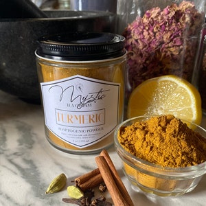 May include: A glass jar of turmeric powder labeled "Mystic Tea Company Turmeric Adaptogenic Powder 4 oz | 118 ml". The jar is sitting on a white marble surface with cinnamon sticks, cloves, and cardamom pods.
