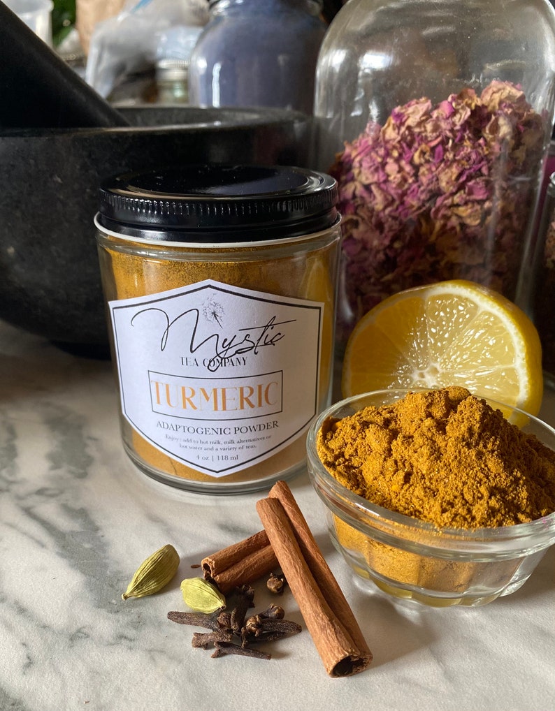 May include: A glass jar of turmeric powder labeled "Mystic Tea Company Turmeric Adaptogenic Powder 4 oz | 118 ml". The jar is sitting on a white marble surface with cinnamon sticks, cloves, and cardamom pods.