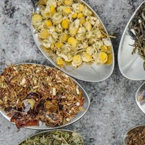 Op de afbeelding: Vier zilveren lepels gevuld met verschillende soorten gedroogde theebladeren. De lepels zijn in een halve cirkel op een grijs, getextureerd oppervlak gerangschikt. De theebladeren omvatten kamille, rooibos, groene thee en zwarte thee.