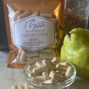 Op de afbeelding: Een glazen kom gevuld met lichtbruine capsules. De kom staat op een wit oppervlak. Op de achtergrond is een gouden zak te zien met de tekst "Mystic Tea Co & Apothecary Ashwagandha Root 100% Organic Herbal Supplement 500mg capsules".