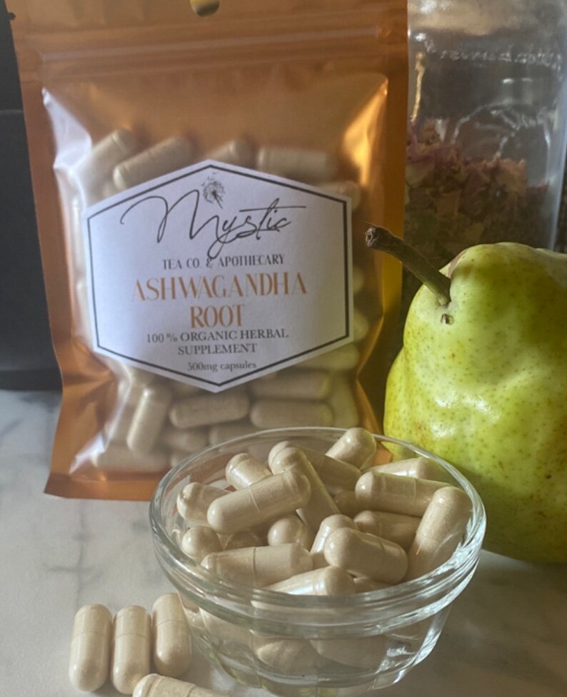 Op de afbeelding: Een glazen kom gevuld met lichtbruine capsules. De kom staat op een wit oppervlak. Op de achtergrond is een gouden zak te zien met de tekst "Mystic Tea Co & Apothecary Ashwagandha Root 100% Organic Herbal Supplement 500mg capsules".