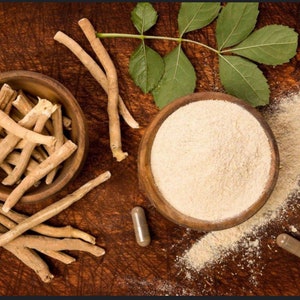 Op de afbeelding: Een close-up van een houten kom gevuld met gedroogde, lichtbruine, wortelvormige stukken Ashwagandha. Een tweede houten kom bevat een lichtbruin poeder. Er zijn ook drie capsules met Ashwagandha-poeder.