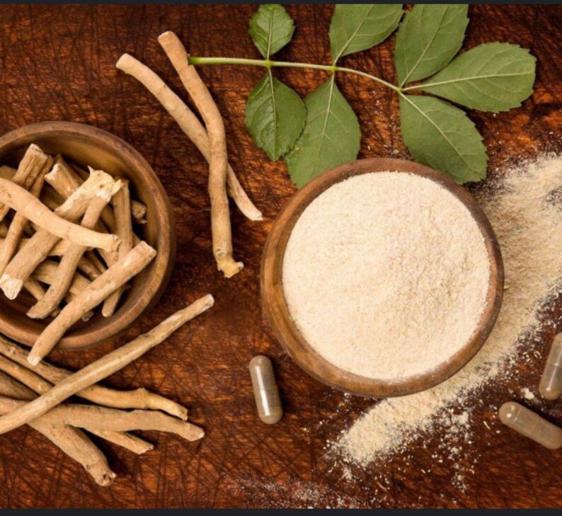 Op de afbeelding: Een close-up van een houten kom gevuld met gedroogde, lichtbruine, wortelvormige stukken Ashwagandha. Een tweede houten kom bevat een lichtbruin poeder. Er zijn ook drie capsules met Ashwagandha-poeder.