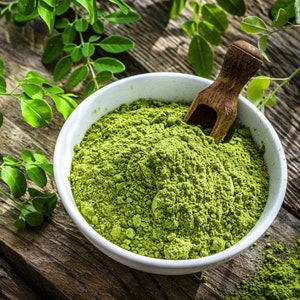 May include: A white bowl filled with green moringa powder, with a wooden scoop resting on top. Fresh moringa leaves are scattered around the bowl.