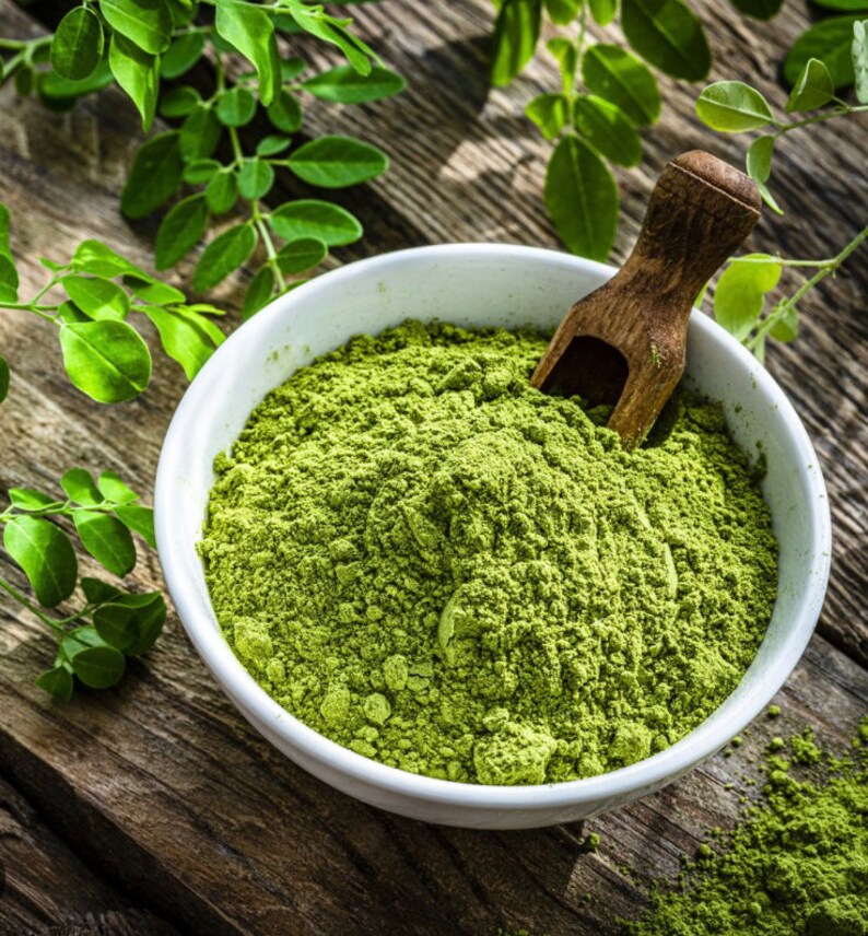 May include: A white bowl filled with green moringa powder, with a wooden scoop resting on top. Fresh moringa leaves are scattered around the bowl.