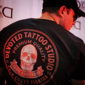 May include: Black t-shirt with a red circle logo that says "Devoted Tattoo Studio" with a skull graphic and the text "Premium Quality 100% Handmade Palm Coast Florida 20 13"