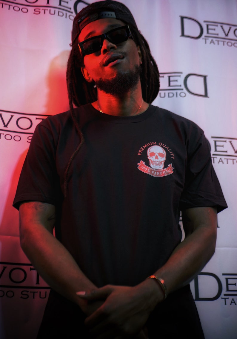 May include: A man wearing a black t-shirt with a white skull graphic and the text "Premium Quality 100% Handmade". He is wearing a black baseball cap and sunglasses.