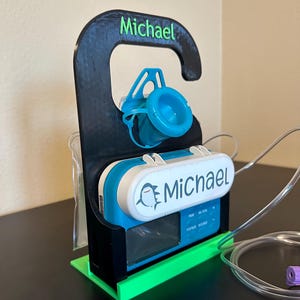 May include: A black 3D printed holder with a green base, designed to hold a medical pump. The pump is blue and white with a shark graphic and the name "Michael" printed on it. The holder has a hook at the top with the name "Michael" printed on it.