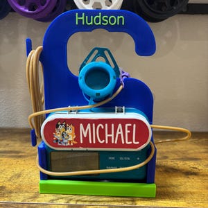May include: A blue and green 3D-printed alarm clock with the name "Hudson" in green. The clock features a red and white label with the name "MICHAEL" and a cartoon image. A tan cord wraps around the clock.