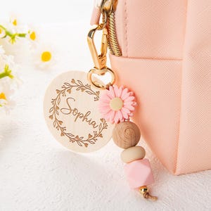 Puede incluir: Un llavero de madera con el nombre "Sophia" grabado en una corona floral. Está unido a un bolso rosa con cremallera dorada. Una flor rosa y cuentas de madera cuelgan del llavero.