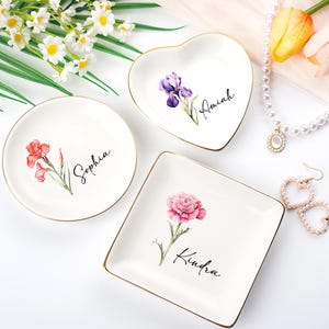 Könnte beinhalten: Drei weiße Keramik-Schmuckschalen mit goldenen Rändern. Eine ist rund, eine herzförmig und eine quadratisch. Jede hat ein Blumendesign und einen Namen: Sophia, Amiah und Kindra. Eine Perlenkette und Ohrringe sind ebenfalls vorhanden.