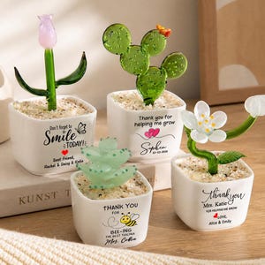 May include: Four small white square ceramic pots containing glass plants: a pink tulip, a green cactus, a light green succulent, and a white flower. Each pot features printed text and graphics, including messages like Thank you for helping me grow.