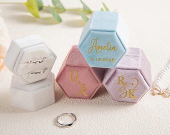 May include: A collection of hexagonal velvet ring boxes in pastel colors: white, light blue, pink, and lavender. Gold lettering personalizes each box with names and dates. A silver ring sits near the boxes. The boxes are designed for engagement rings.