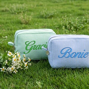 May include: Two small, rectangular cosmetic bags with zipper closures rest on green grass. One is light green with "Gracie" embroidered in green, the other is light blue with "Bonie" embroidered in blue. A small bouquet of white flowers is in the foreground.