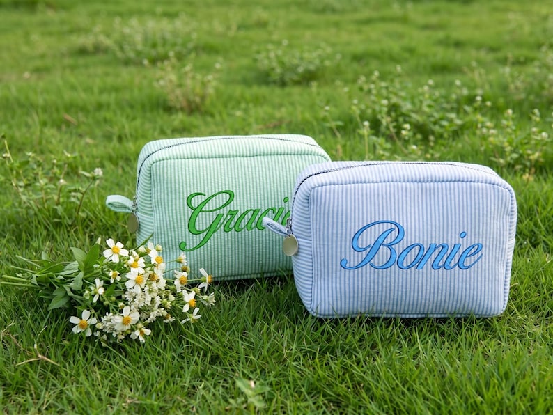 May include: Two small, rectangular cosmetic bags with zipper closures rest on green grass. One is light green with "Gracie" embroidered in green, the other is light blue with "Bonie" embroidered in blue. A small bouquet of white flowers is in the foreground.