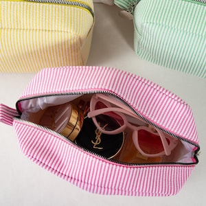May include: Pink and white striped cosmetic bag with a black zipper, open to reveal sunglasses and makeup. Two other bags, one yellow and one green, are visible in the background. The bags have a seersucker texture.