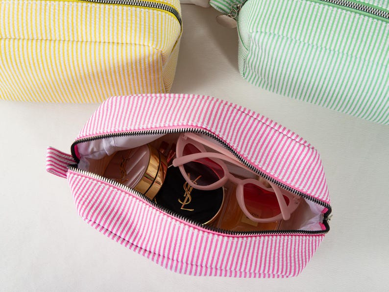 May include: Pink and white striped cosmetic bag with a black zipper, open to reveal sunglasses and makeup. Two other bags, one yellow and one green, are visible in the background. The bags have a seersucker texture.