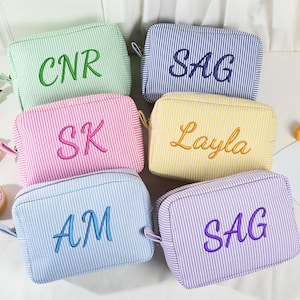 May include: Six striped cosmetic bags in various colors, including green, blue, pink, yellow, and purple. Each bag has embroidered lettering: CNR, SAG, SK, Layla, AM, and SAG. The bags have a rectangular shape with a zipper closure.
