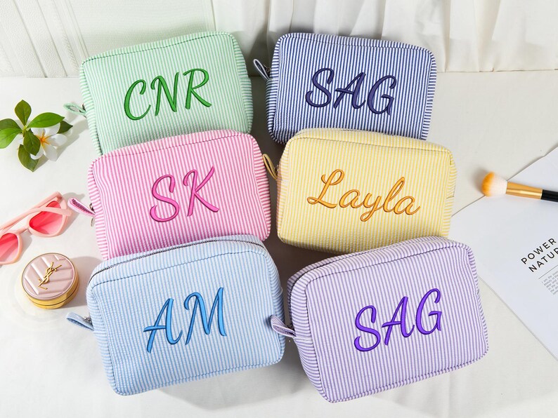 May include: Six striped cosmetic bags in various colors, including green, blue, pink, yellow, and purple. Each bag has embroidered lettering: CNR, SAG, SK, Layla, AM, and SAG. The bags have a rectangular shape with a zipper closure.