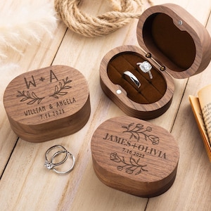 以下が含まれることがあります： 名前と日付が刻まれた楕円形の木製リングボックス。ダイヤモンド付きのシルバーリングが入っています。ボックスは濃い茶色です。1つのボックスが開いており、リングが見えます。ボックスのテキストは「W+A WILLIAM & ANGELA 7.16.2025」と「JAMES+OLIVIA 7.16.2025」です。