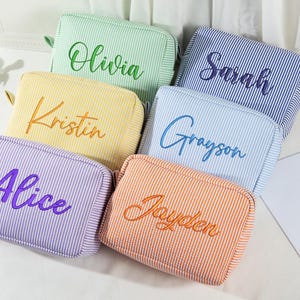 May include: Six colorful, striped cosmetic bags with embroidered names: Olivia, Sarah, Alice, Kristin, Grayson, and Jayden. The bags are in shades of green, blue, purple, yellow, orange, and blue. A makeup brush and sunglasses are visible.
