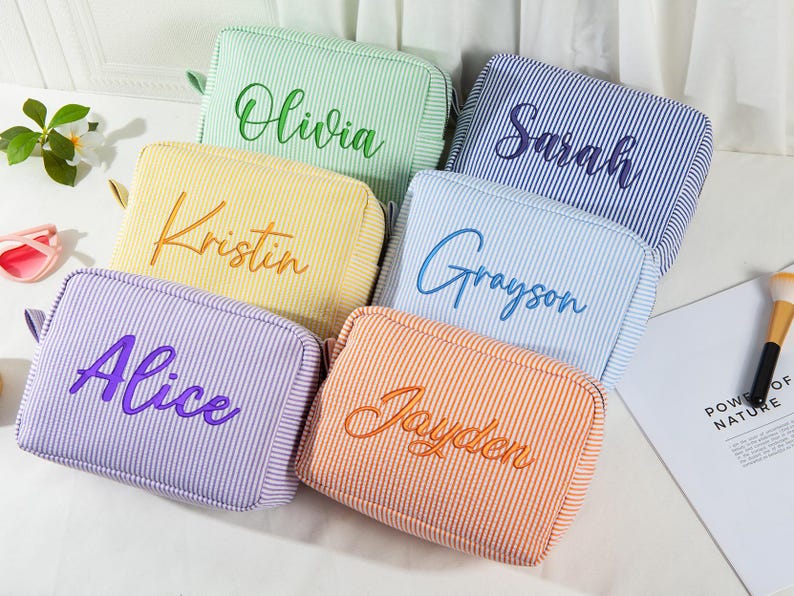 May include: Six colorful, striped cosmetic bags with embroidered names: Olivia, Sarah, Alice, Kristin, Grayson, and Jayden. The bags are in shades of green, blue, purple, yellow, orange, and blue. A makeup brush and sunglasses are visible.