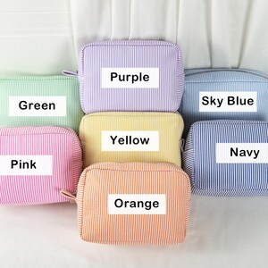 May include: A collection of seven rectangular cosmetic bags in various colors, including green, purple, sky blue, yellow, pink, orange, and navy. Each bag features a striped pattern and a label with its color name.