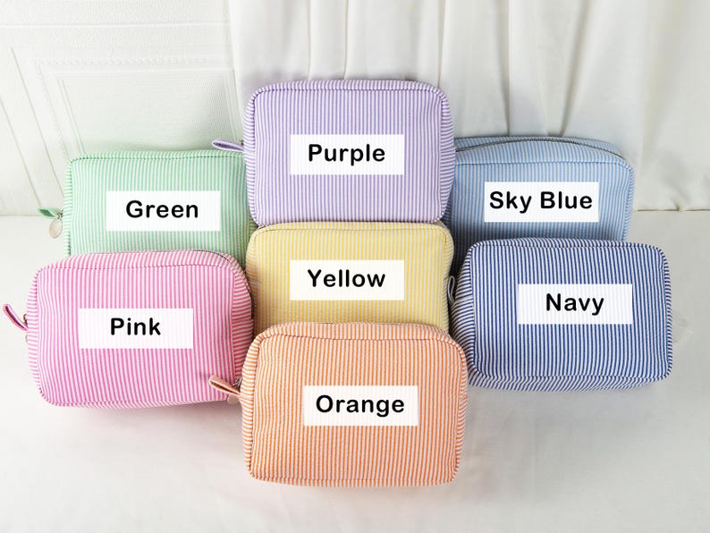 May include: A collection of seven rectangular cosmetic bags in various colors, including green, purple, sky blue, yellow, pink, orange, and navy. Each bag features a striped pattern and a label with its color name.
