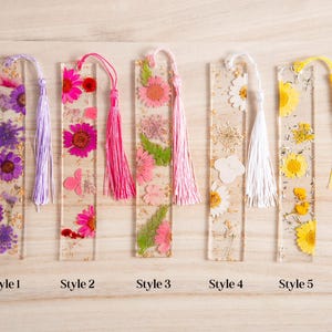 May include: Five rectangular resin bookmarks with dried flowers and tassels. Styles include purple, pink, and yellow flowers, with matching tassels. Each bookmark is labeled with a style number.