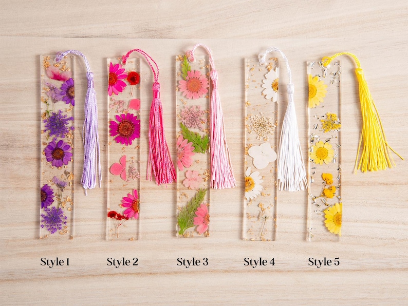 May include: Five rectangular resin bookmarks with dried flowers and tassels. Styles include purple, pink, and yellow flowers, with matching tassels. Each bookmark is labeled with a style number.