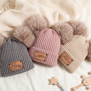 Könnte beinhalten: Drei gerippte Strickmützen in Grau, Rosa und Beige, jede mit einem Kunstfell-Bommel und einem Namensschild aus Kunstleder. Die Namen "Elias", "Sophia" und "Victoria" sind abgebildet.