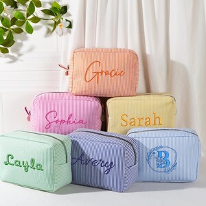 May include: A collection of six personalized cosmetic bags in various colors, including orange, pink, yellow, green, blue, and navy. Each bag features a vertical stripe pattern and embroidered names or initials in complementary colors. The bags are arranged in a stacked display.