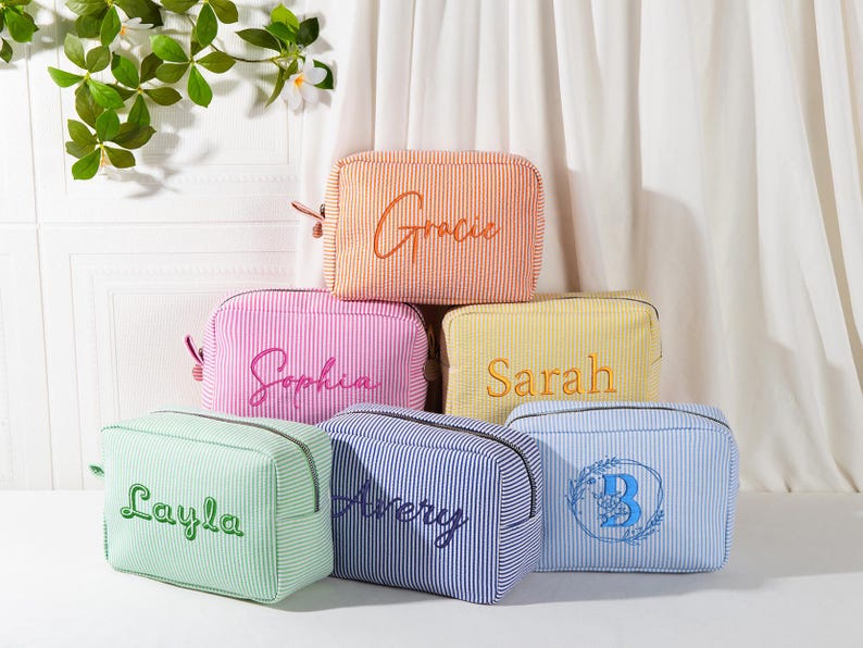 May include: A collection of six personalized cosmetic bags in various colors, including orange, pink, yellow, green, blue, and navy. Each bag features a vertical stripe pattern and embroidered names or initials in complementary colors. The bags are arranged in a stacked display.
