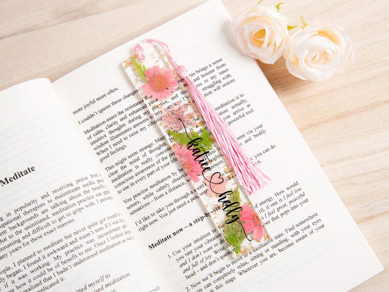 May include: A clear acrylic bookmark with pink flowers and green leaves, with the names "Katie" and "Holly" in black script. The bookmark has a pink tassel and is placed on an open book.