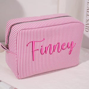 May include: A pink and white striped cosmetic bag with the name "Finney" embroidered in a cursive font. The bag has a zipper closure and a small loop on the side. The bag is rectangular and appears to be made of fabric.