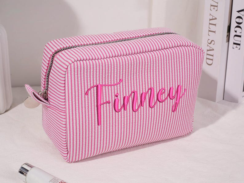 May include: A pink and white striped cosmetic bag with the name "Finney" embroidered in a cursive font. The bag has a zipper closure and a small loop on the side. The bag is rectangular and appears to be made of fabric.
