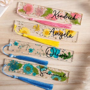 May include: Clear resin bookmarks with dried flowers and personalized names. The bookmarks feature pink, yellow, and blue flowers, with names like "Kendrick", "Angela", "Olivia", and "Samuel" in black script. Each bookmark has a tassel.