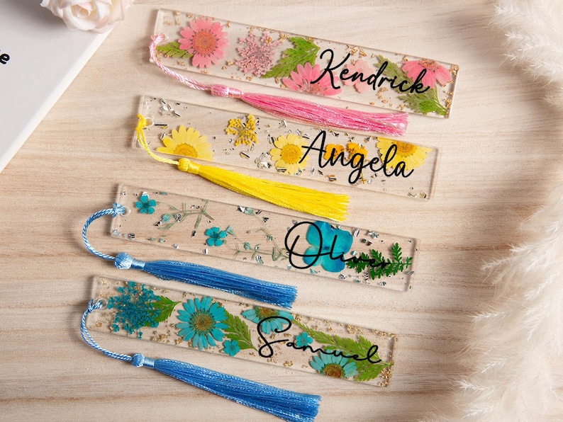 May include: Clear resin bookmarks with dried flowers and personalized names. The bookmarks feature pink, yellow, and blue flowers, with names like "Kendrick", "Angela", "Olivia", and "Samuel" in black script. Each bookmark has a tassel.