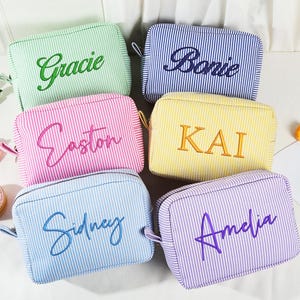 May include: Six personalized cosmetic bags in various colors, including green, blue, pink, yellow, and purple. Each bag features a vertical striped pattern and a name embroidered in a complementary color. The bags have a zipper closure and a small handle.