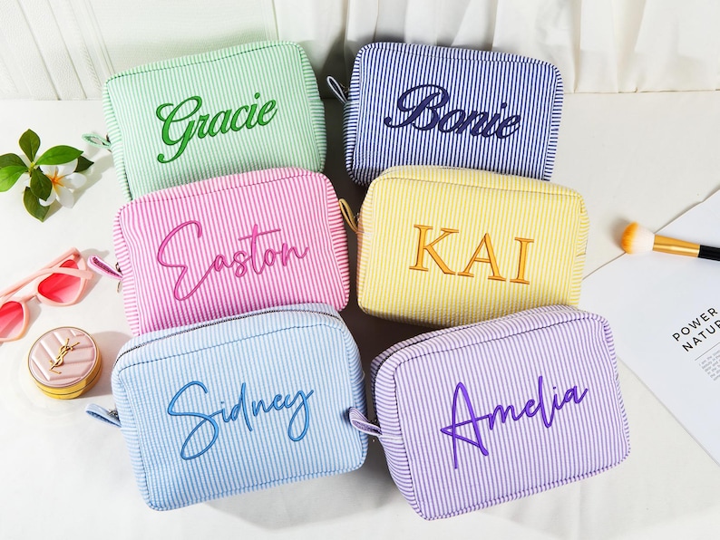 May include: Six personalized cosmetic bags in various colors, including green, blue, pink, yellow, and purple. Each bag features a vertical striped pattern and a name embroidered in a complementary color. The bags have a zipper closure and a small handle.